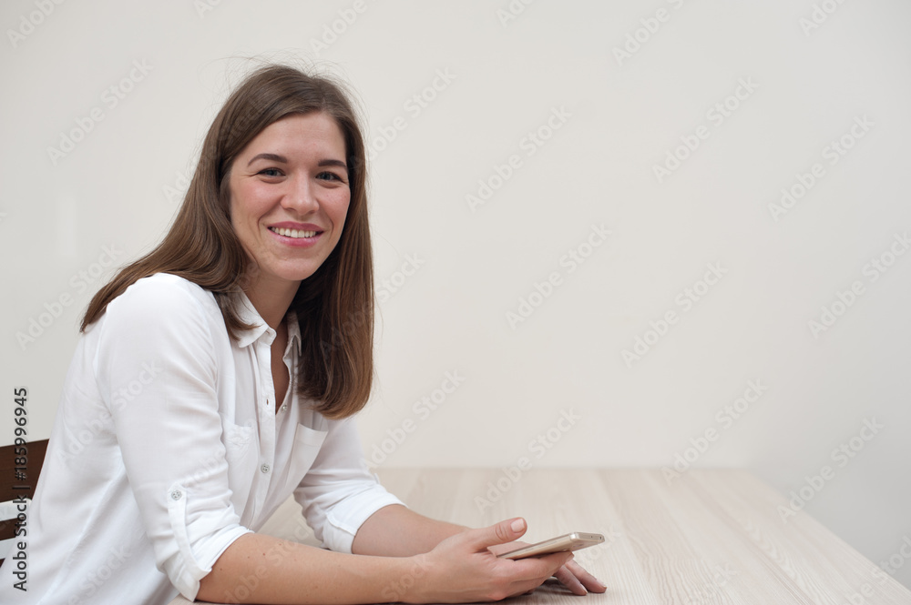 Portrait of beautiful positive young woman sitting at the table with smartphone on white wall background. Copyspace Portrait of beautiful positive young woman sitting at the table with smartphone on white wall background. Copyspace
