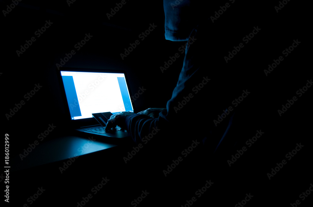 Φωτογραφία Credit card thief wearing hood, sitting in the front of laptop, credit cards, in a dark room, working with the password, conceptual image of internet fraud