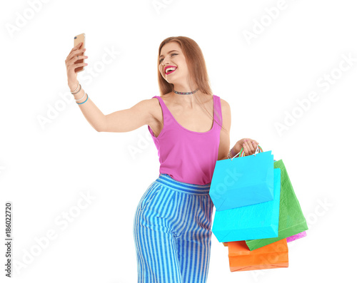 Happy young woman with shopping bags taking selfie on white background
