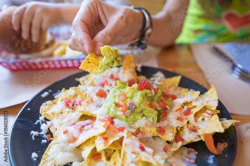 woman hand dipping nacho chips in avocado guacamole, cheese and chopped tomato on black plate, on wooden table with paper placemats at restaurant
