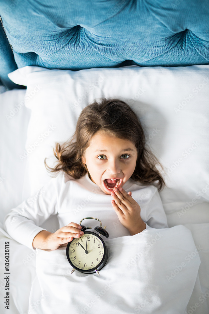 Top view of cheerful girl lying in bed with alarm clock at home, looking at camera