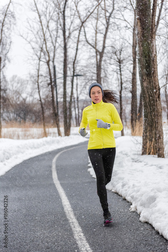 Winter runner girl training running in snow wearing cold weather clothes, gloves, warm yellow jacket. Asian woman jogging in park during snowfall. Active and fitness lifestyle. © Maridav