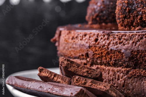 Chocolate biscuit cake decorated with chocolate glaze and crumbs on a white plate on a dark background. Close-up