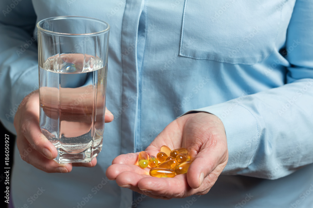 Yellow medication capsules of omega 3, fish oil, healthy supplement pills in the woman palm hand and glass with water in the other hand Yellow medication capsules of omega 3, fish oil, healthy supplement pills in the woman palm hand and glass with water in the other hand