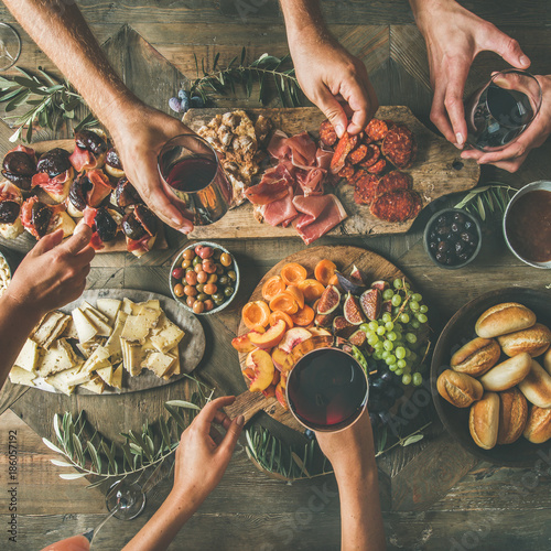 Flat-lay of friends hands eating and drinking together. Top view of people having party, celebrating together at vintage wooden rustic table set with different wine snacks and fingerfoods, square crop