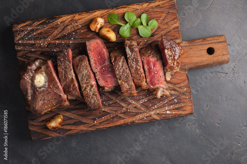 Closeup ready to eat steak new York beef breeds of black Angus with herbs, garlic and butter on a wooden Board. The finished dish for dinner on a dark stone background. Top view photo