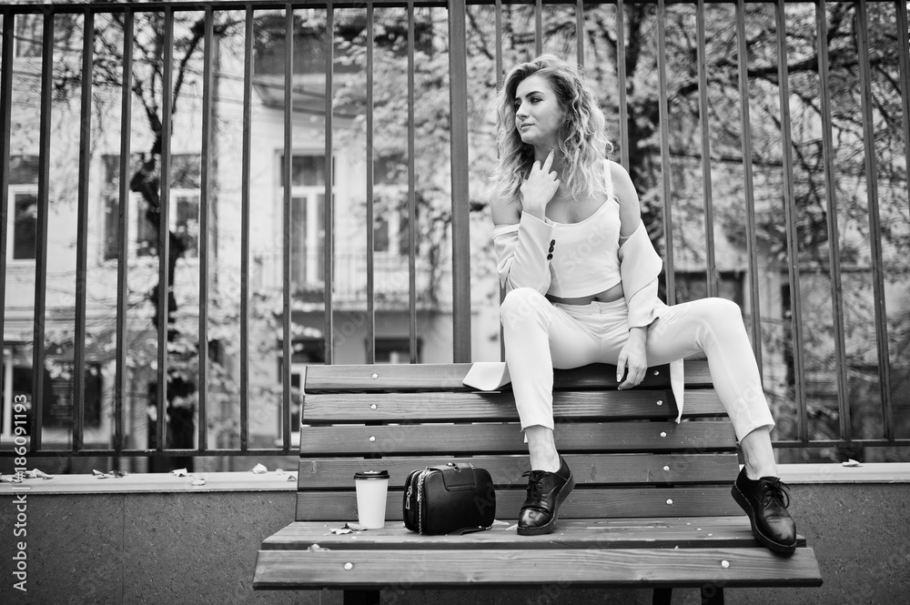 Stylish curly blonde model girl wear on white with cup of coffee posing at bench at fall.
