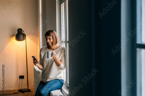 Pretty teenage girl sitting on windowsill with smartphone and texting.