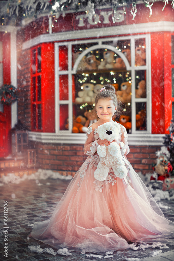 Beautiful little baby girl in a dress holding  teddy bear, on the background of the showcase red with toys