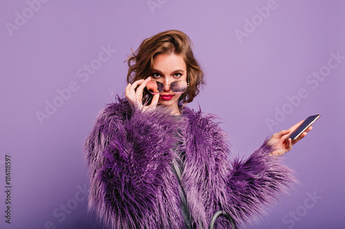 Foto Interested girl in stylish purple fur coat looking to camera holding sparkle sunglasses