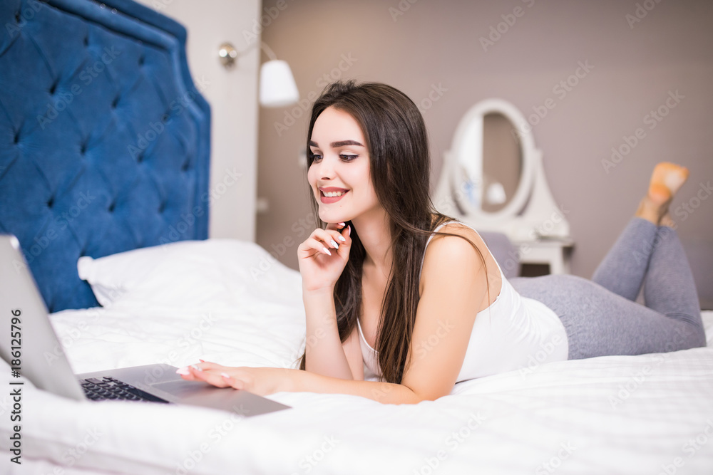 Pretty Smiling Woman Busy Browsing on Laptop While Having a Cup of Coffee on White Bed. Pretty Smiling Woman Busy Browsing on Laptop While Having a Cup of Coffee on White Bed.