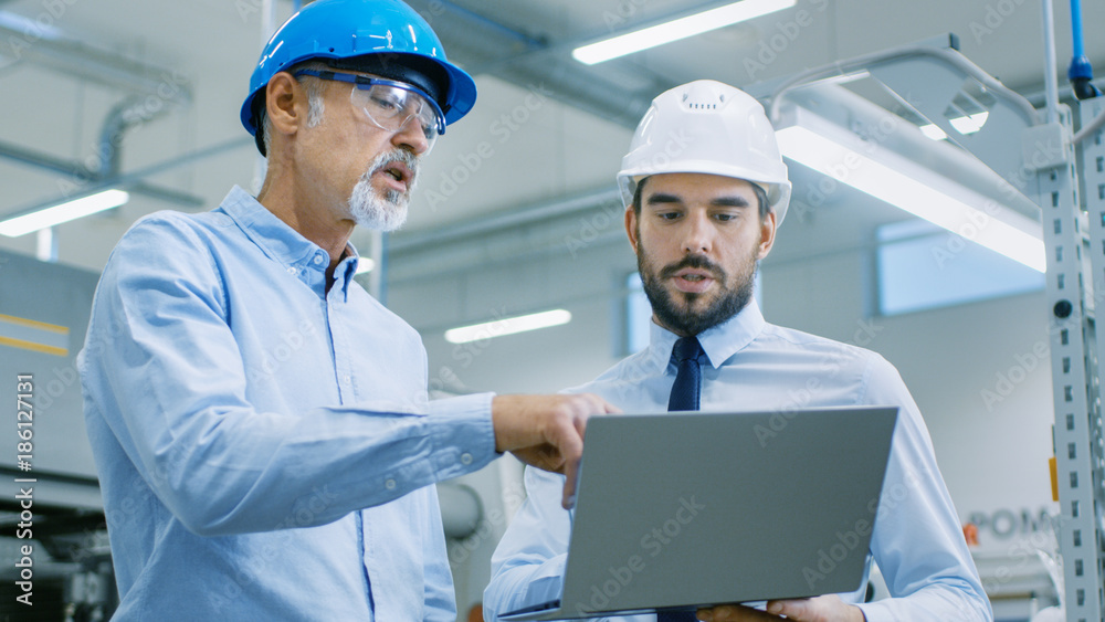 Head of the Department Holds Laptop and Discusses Product Details with Chief Engineer. They Wear Hard Hats and Work at the Modern Factory. Head of the Department Holds Laptop and Discusses Product Details with Chief Engineer. They Wear Hard Hats and Work at the Modern Factory.