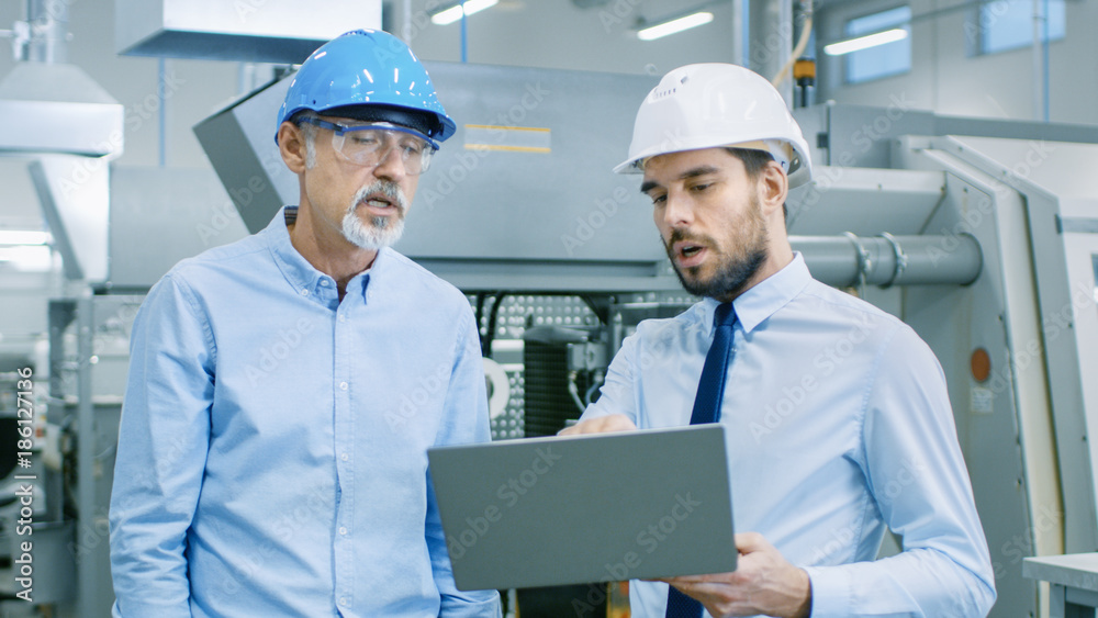 Head of the Department Holds Laptop and Discusses Product Details with Chief Engineer. They Wear Hard Hats and Work at the Modern Factory. Head of the Department Holds Laptop and Discusses Product Details with Chief Engineer. They Wear Hard Hats and Work at the Modern Factory.
