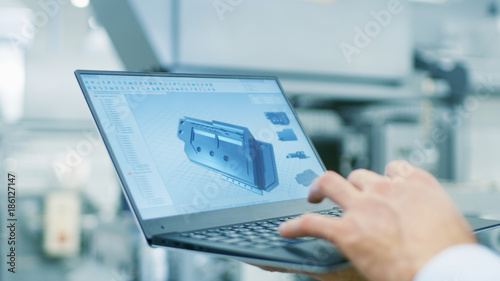 Close-up of the Engineer Holding Laptop with CAD Component Model on Screen. In the Background Modern Factory Equipment.