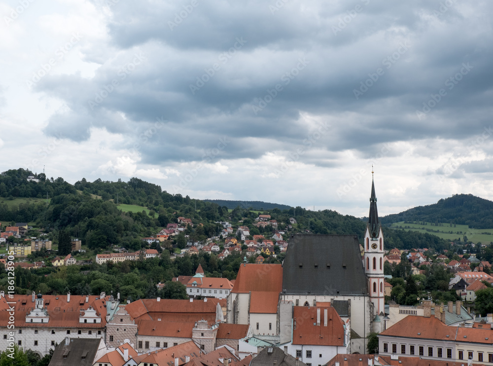 View of the charming medieval town, Cesky Krumlov, from the castle, view of St Vitus Chruch (Kostel vs Vitas) and the red roofed village surrounding the church. Stormy clouds and green hills.