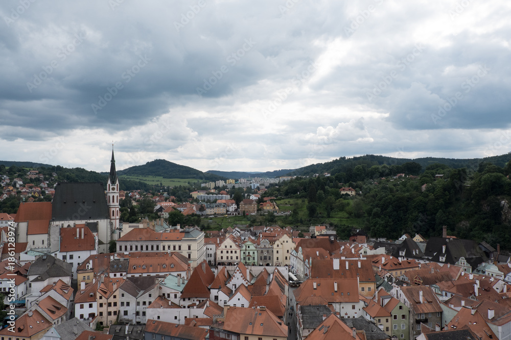View of the charming medieval town, Cesky Krumlov, from the castle tower. Cityscape of Cesky Krumlov with red roof houses, green mountains, stormy sky, and the Church of St Vitus (Kostel sv Vita).