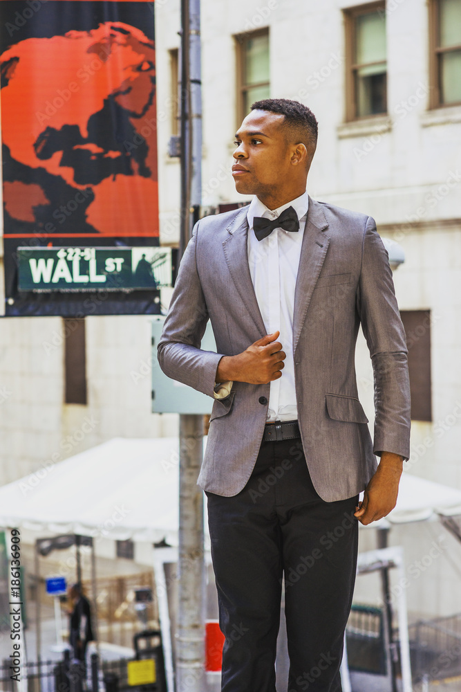 African American Businessman working in New York. Wearing gray blazer, bow  tie, black pants, young black college student standing on Wall Street  outside office, looking away, confident, successful. . Photos | Adobe Stock