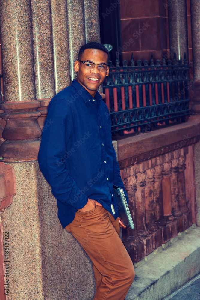African American college student studying in New York, wearing blue shirt, brown  pants, glasses, holding laptop computer, standing against vintage style  wall on street, smiling, waiting for you. Stock Photo | Adobe