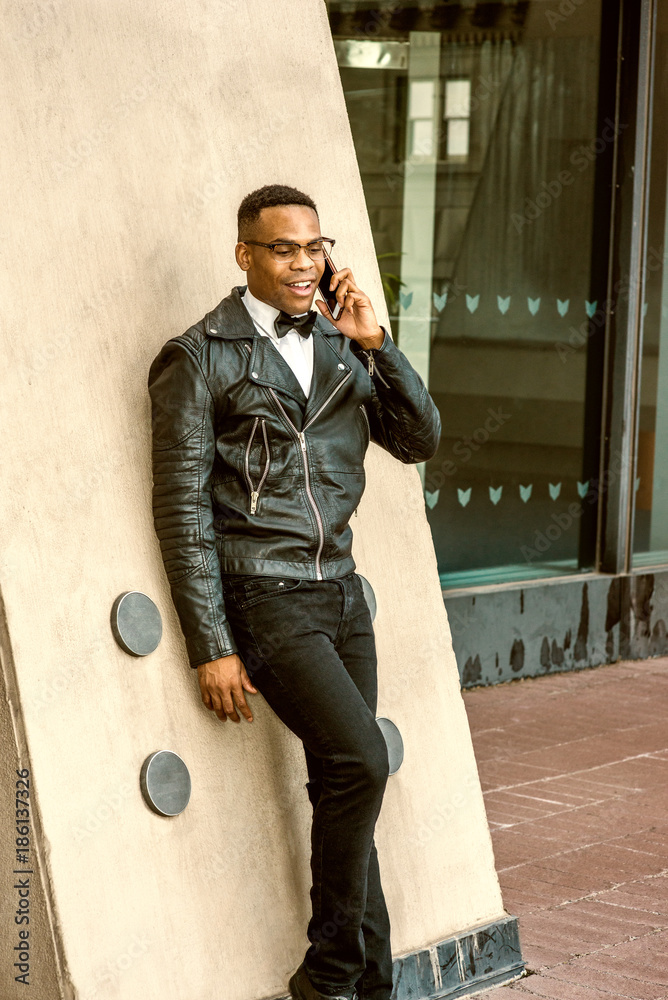 Man Urban Autumn/Spring Fashion. Wearing black leather jacket, white  undershirt, black bow tie, jeans, glasses, African American guy standing  against wall on street in New York, talking on cell phone Stock Photo