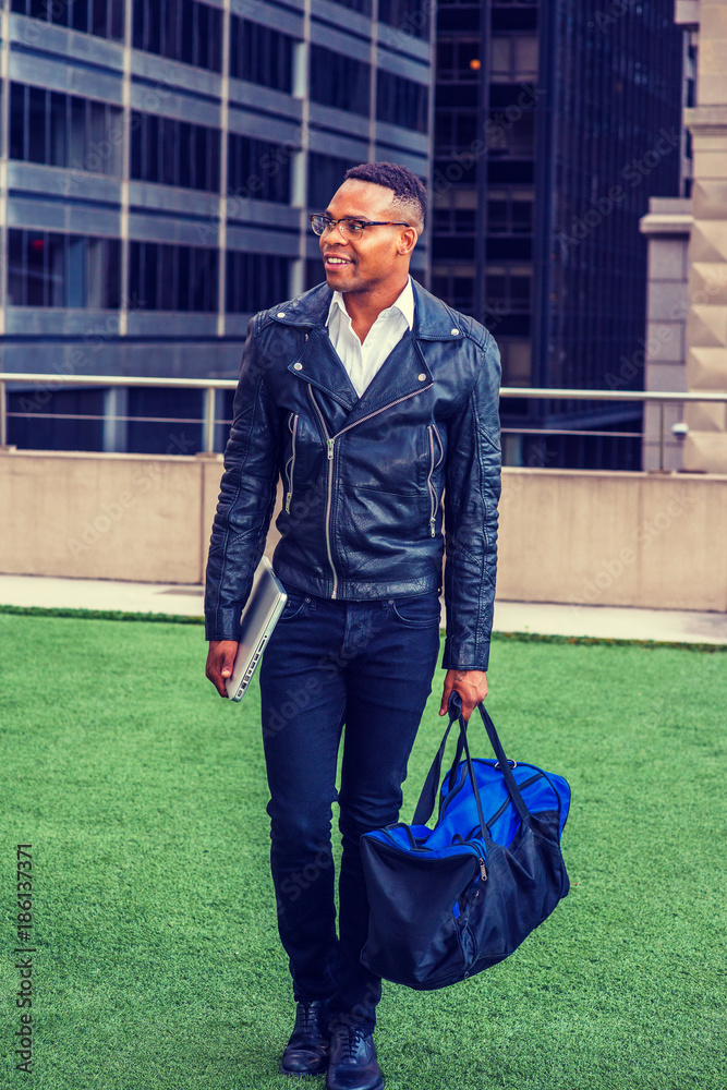 Man Urban Casual Fashion. Wearing black leather jacket, jeans, leather  shoes, glasses, holding laptop computer, carrying duffel bag, African  American college student walking on campus in New York. Stock Photo | Adobe