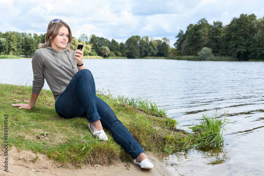 Fototapeta premium Blond woman sitting on river bank with mobile phone in hand, looking at screen