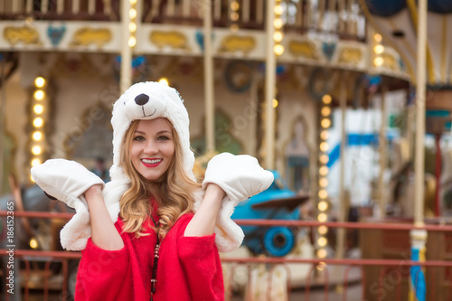 Emotional young woman wearing red knitted sweater and funny hat, posing at the background of carousel with lights