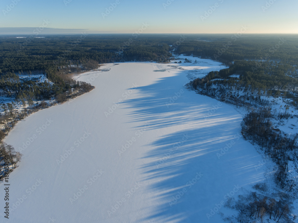 Aerial view over frozen lake cover in white snow with long blue shadows from trees in Latazeris, Lithuania. During cold winter day.