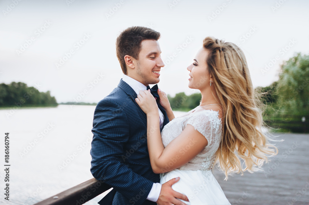 Beautiful and young bride and groom tenderly embrace on the pier by the river. Attractive bride with long blond hair. Wedding ceremony in nature by the river.
