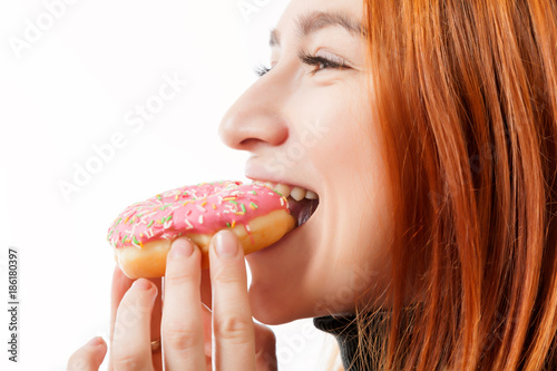 A young red-haired woman in a black turtleneck and a kitchen apron laughs and eats a donut pink with pleasure on a white isolated background