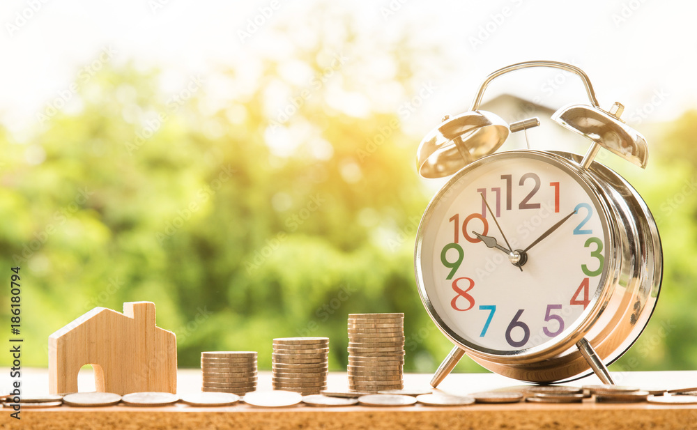 alarm clock and wooden house model and step of coins stacks with nature background, money, saving and investment or family planning concept, over sun flare silhouette tone.