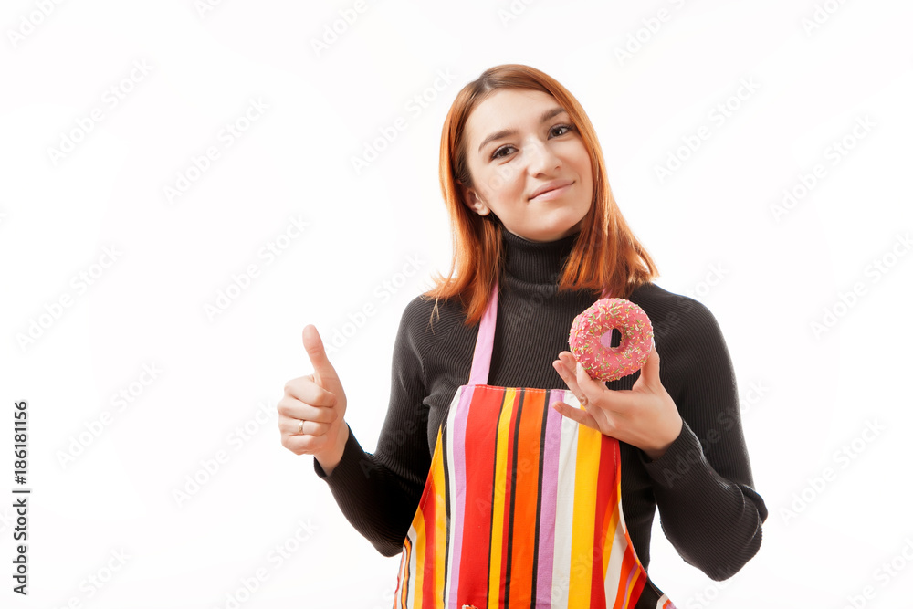 Young red-haired woman in black sweater and kitchen apron holds a pink donut with chocolate filling in her hand, shows it to the camera and smiles on a white isolated background Young red-haired woman in black sweater and kitchen apron holds a pink donut with chocolate filling in her hand, shows it to the camera and smiles on a white isolated background