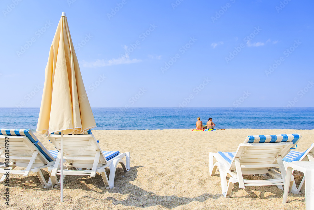 Sunbathing on resort beach on hot, sunny day. Umbrellas and sunbeds on white sand against the blue sea and clear sky. Two women sit on sand and relax near sea.