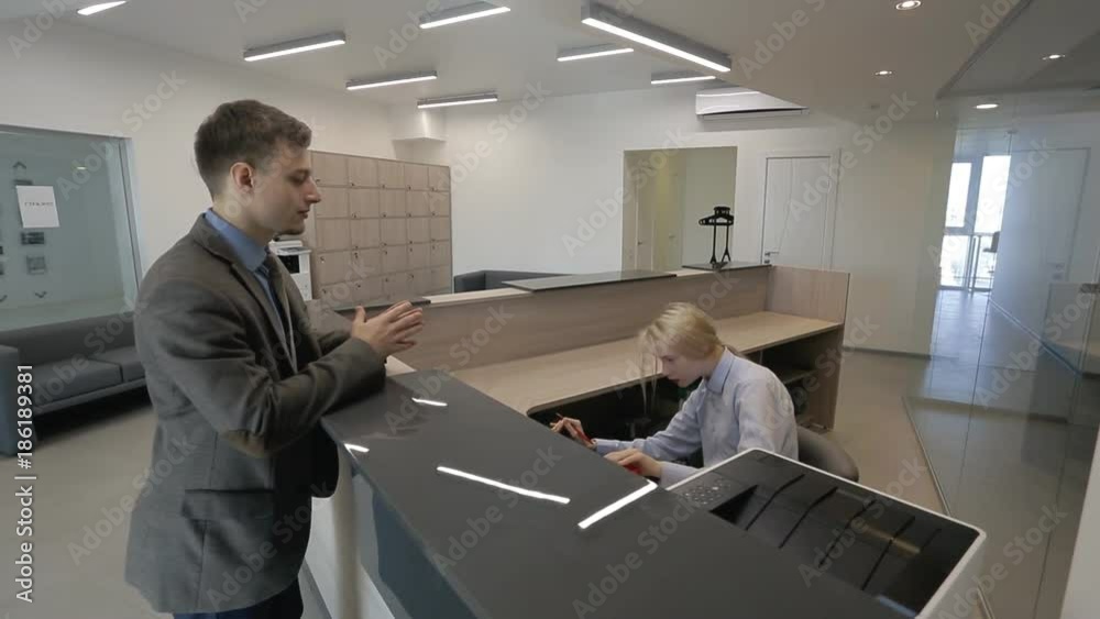Young employees are talking at reception desk of company indoors. Male specialist communicates with woman receptionist in modern office. Handsome man in business suit is standing at high table and