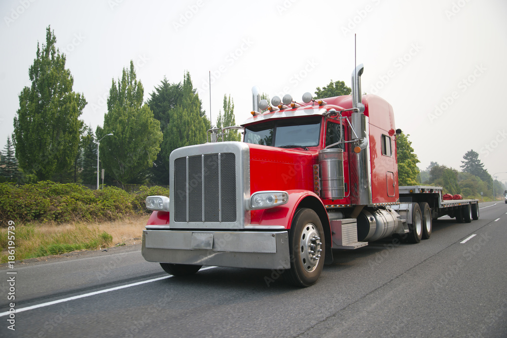 Powerful red big rig semi truck with flat bed step down semi trailer move on smog road for loading cargo