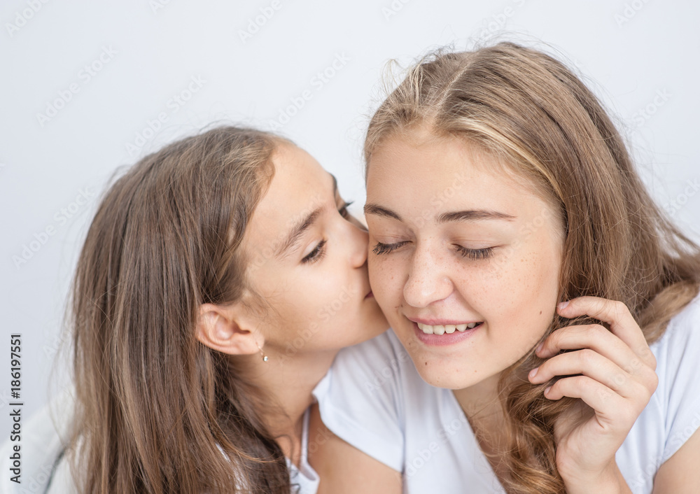 girl is  kissing her beautiful young mom in cheek while sitting on bed at home