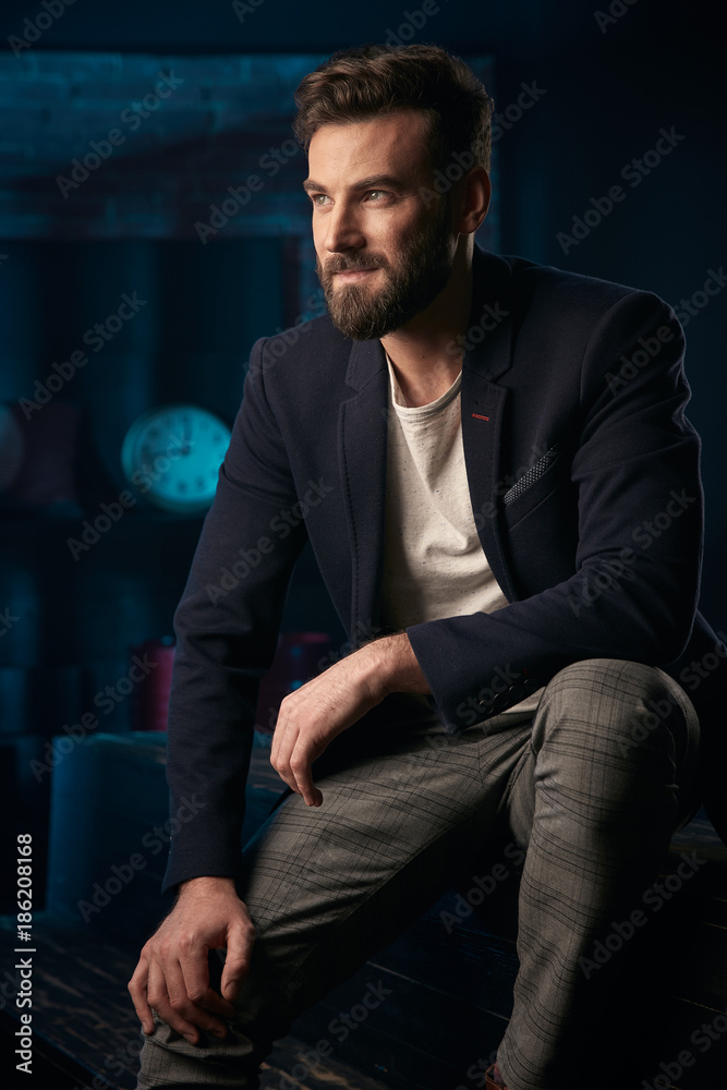 Studio portrait of serious handsome man with dark hair, beard and mustache wearing dark blue jacket, gray pants sitting on wooden surface. Blue lighted brick background with clock and barrel Studio portrait of serious handsome man with dark hair, beard and mustache wearing dark blue jacket, gray pants sitting on wooden surface. Blue lighted brick background with clock and barrel