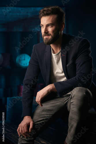 Studio portrait of serious handsome man with dark hair, beard and mustache wearing dark blue jacket, gray pants sitting on wooden surface. Blue lighted brick background with clock and barrel