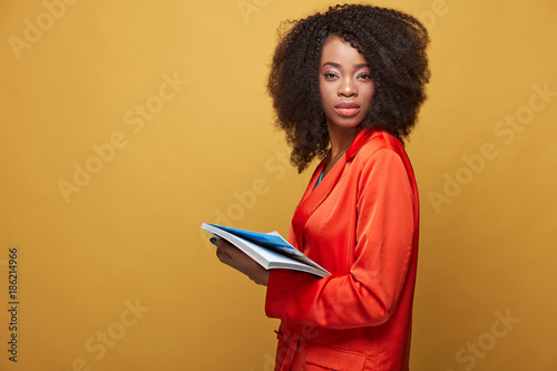 Colorful portrait of young african girl with afro hairstyle. Serious girl wearing orange jacket holds copybook in her hand and posing on yellow background. Studio shot.