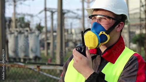Electrician in respirator with portable radio transmitter on electric power station. Power engineering specialist in gas mask, goggles and hard hat on heat power plant communicates via radio station