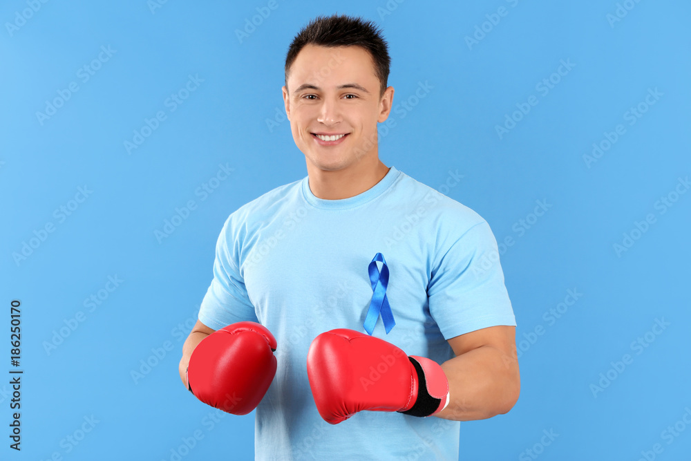 Φωτογραφία Young man in t-shirt with blue ribbon wearing boxing gloves on color background