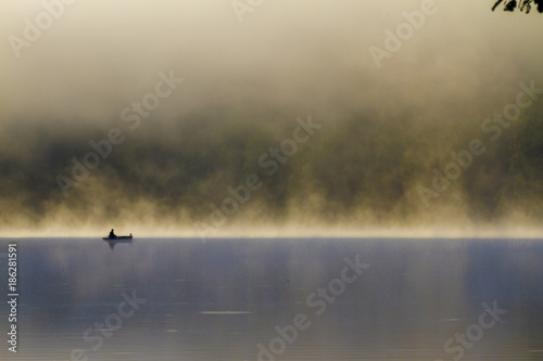Fisherman on a wood boat in a lake at sunrise in a foggy day