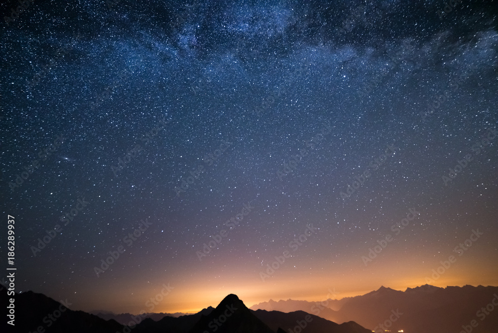 Fototapeta premium The wonderful starry sky on Christmas time and the majestic high mountain range of the Italian Alps, with glowing villages below.
