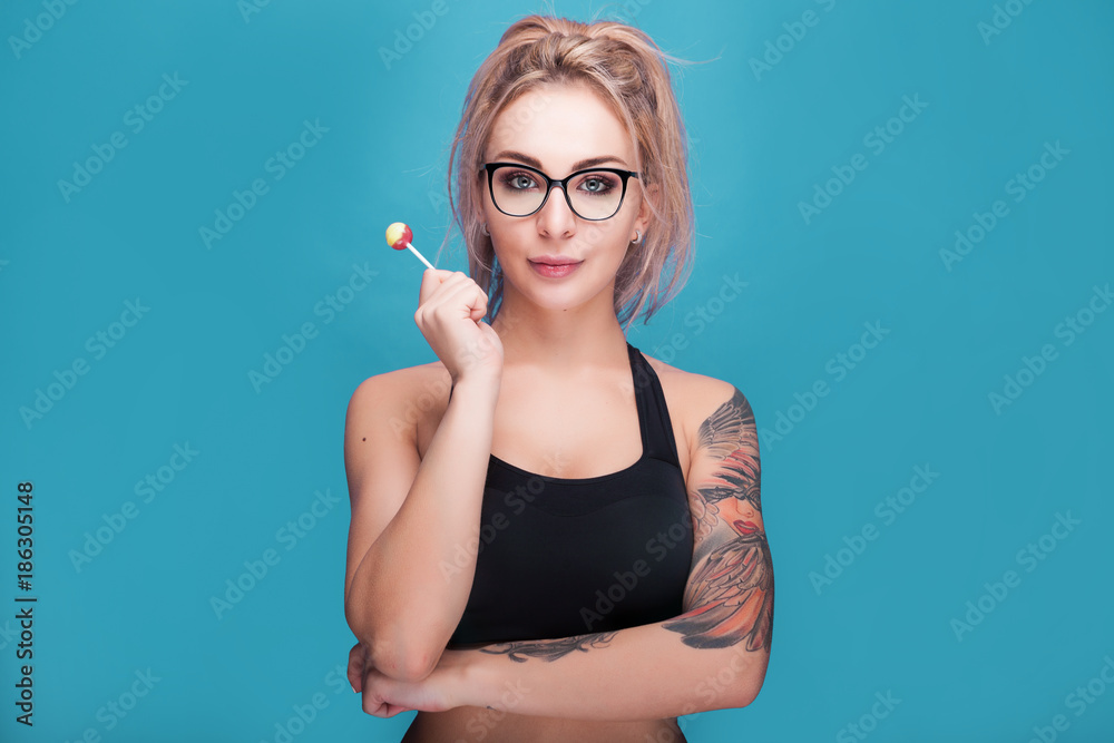 Blonde woman wearing sport outfit and glasses holding a lollipop in hands on blue cyan background Blonde woman wearing sport outfit and glasses holding a lollipop in hands on blue cyan background