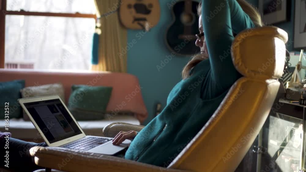 Steadicam clip of profile of mature pretty woman sitting in easy chair lifting her arm and working on computer in a home.