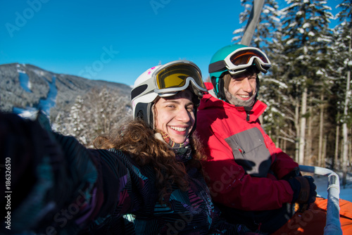 Young pair man and woman of skiers riding up on ski lift, taking selfie, smiling at the camera. Sunny day in the mountains. Close-up copyspace seasonal activity sport hobby recreation travel concept