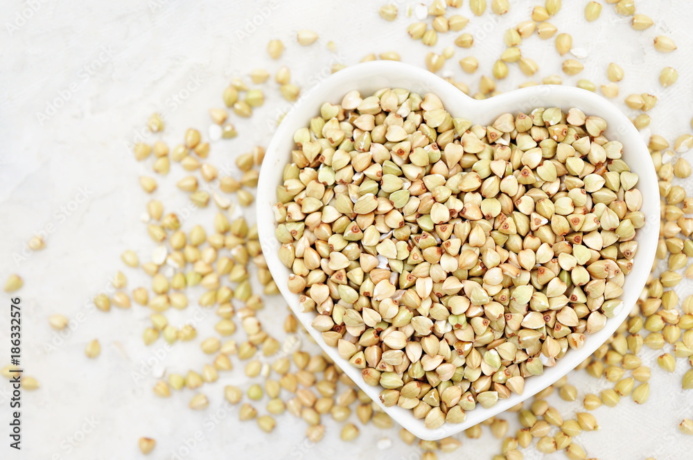 Heart shaped bowl of green buckwheat on buckwheat groat background, gluten free ancient grain for healthy diet, selective focus