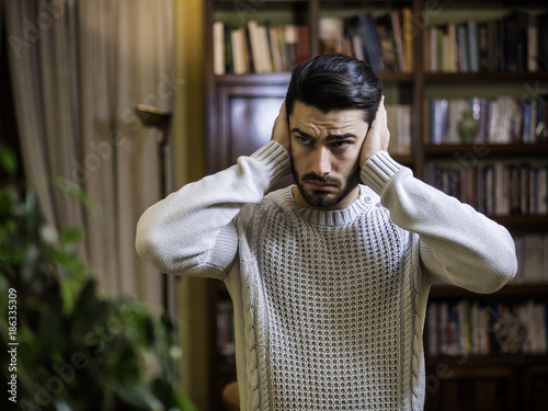 Handsome young man covering his ears, stressed or unhappy because of too much noise. Looking away to a side. Indoors shot
