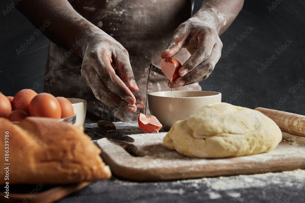 Cropped image of back man in apron, breaks eggs in bowl as prepares filling in buns, surrounded with prepared dough, rolling pin and white loaf, isolated over black background. Handmade concept