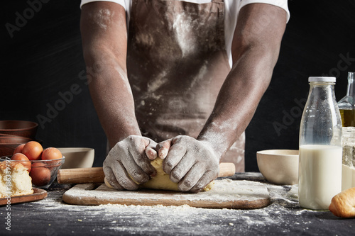 Unrecognizable dark skinned male baker kneads dough, wears apron dirty in flour, surrounded with different ingridients, wants to impress guests with delicious pie, isolated over black background