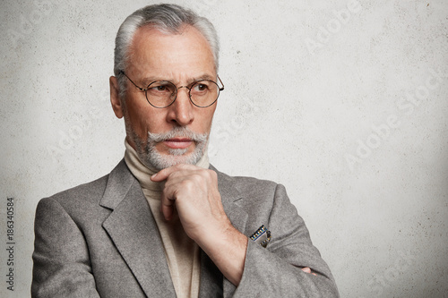 Thoughtful elderly man with grey hair and thick beard, looks pensively aside, thinks about his actions on weekends, going to spend them in family circle. Prosperous male business owner indoor